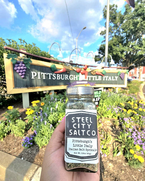Pittsburgh Italian Salt Sprinkle by Steel City Salt Co. held in front of Bloomfield's Little Italy sign, showcasing local flavor and heritage blend.