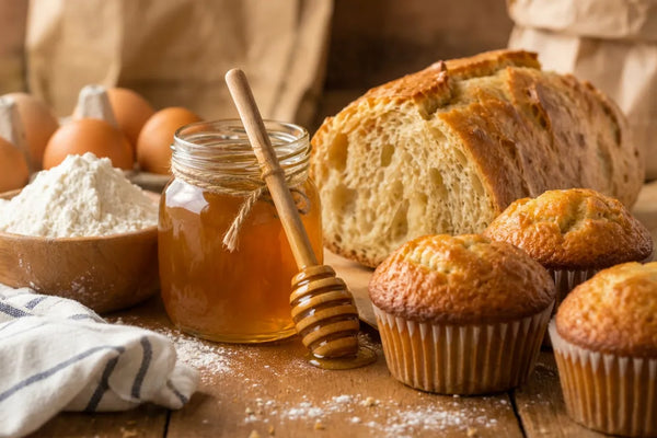 Jar of raw honey with a dipper beside flour and eggs, homemade bread and muffins showing honey's role in baking in a tight close up photo