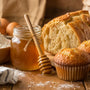 Jar of raw honey with a dipper beside flour and eggs, homemade bread and muffins showing honey's role in baking in a tight close up photo
