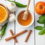 Fresh ingredients for infused honey including sliced oranges, garlic, cinnamon sticks, mint leaves, ginger, honey with dipper, apple, tomato, and green chili on a white wooden background.