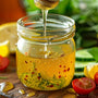 A bright, fresh kitchen scene with a clear glass jar of golden honey vinaigrette dressing on a rustic wooden countertop. A honey dipper drizzles honey into the jar, creating a glistening swirl of light. Surrounding the jar are vibrant salad ingredients—mi