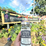 Pittsburgh Italian Salt Sprinkle by Steel City Salt Co. held in front of Bloomfield's Little Italy sign, showcasing local flavor and heritage blend.