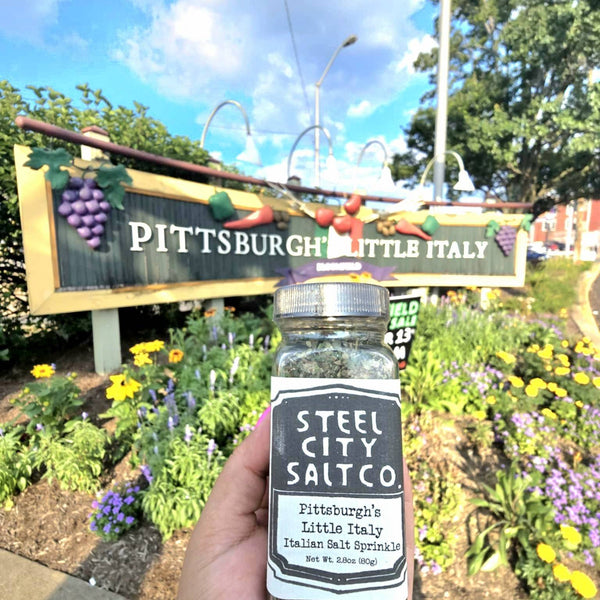 Pittsburgh Italian Salt Sprinkle by Steel City Salt Co. held in front of Bloomfield's Little Italy sign, showcasing local flavor and heritage blend.