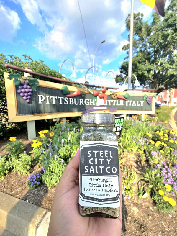 Pittsburgh Italian Salt Sprinkle by Steel City Salt Co. held in front of Bloomfield's Little Italy sign, showcasing local flavor and heritage blend.