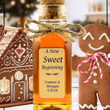 Personalized gingerbread honey bottle with a cork lid, tied with twine, labeled 'A New Sweet Beginning' with names Connor & Morgan and date 6.15.24. Surrounded by festive gingerbread cookies and a decorated gingerbread house.