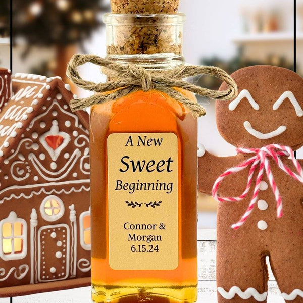 Personalized gingerbread honey bottle with a cork lid, tied with twine, labeled 'A New Sweet Beginning' with names Connor & Morgan and date 6.15.24. Surrounded by festive gingerbread cookies and a decorated gingerbread house.