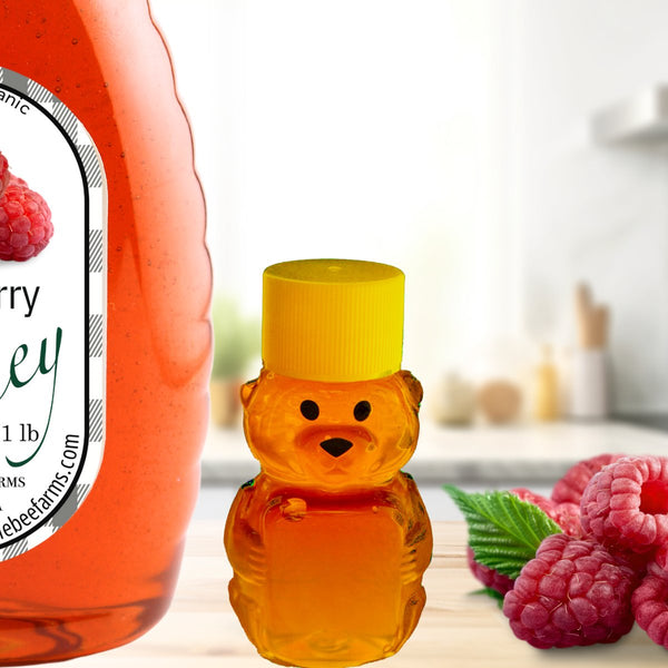 Small bear-shaped honey bottle next to a large raspberry infused honey jar, set in a cozy kitchen with fresh raspberries for a sweet touch.